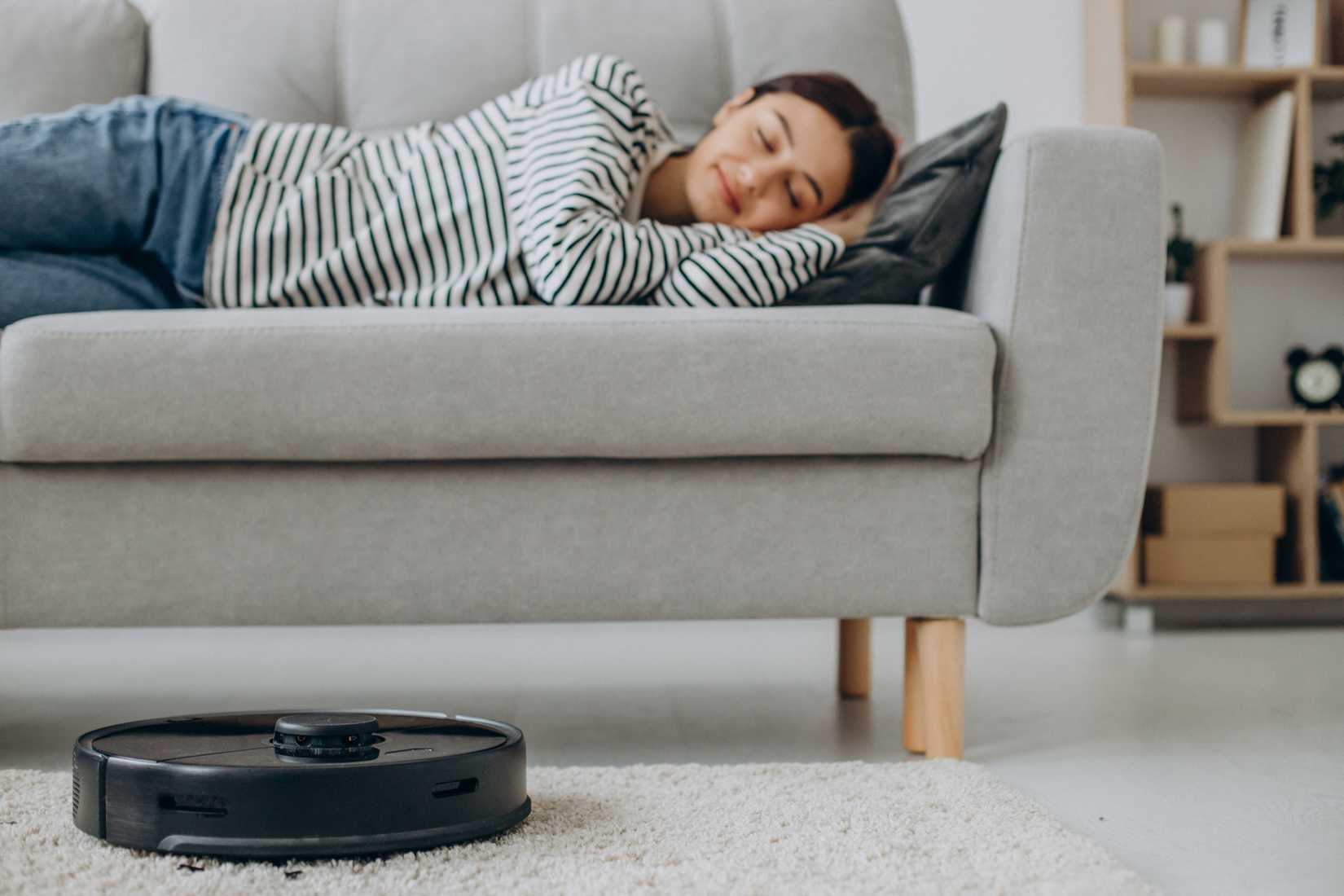 A woman sleeps on the couch while a robot vacuum cleans the carpet.