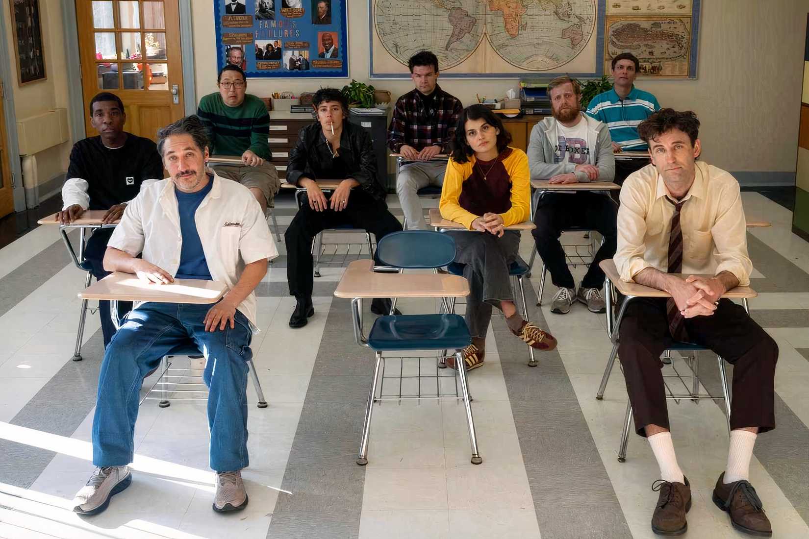 A group of people sit down in desks in Never Change.
