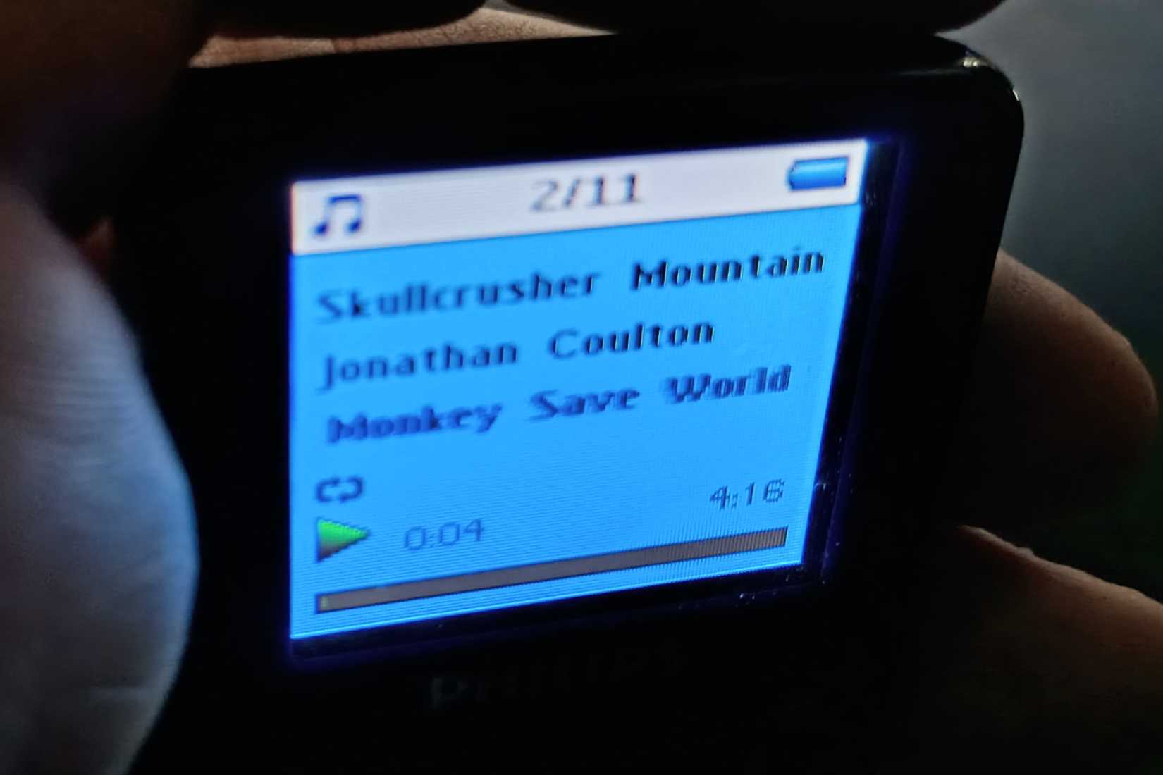 A hand holds a Philips Go Gear MP3 player playing Skull crusher Mountain by Jonathan Coulton.