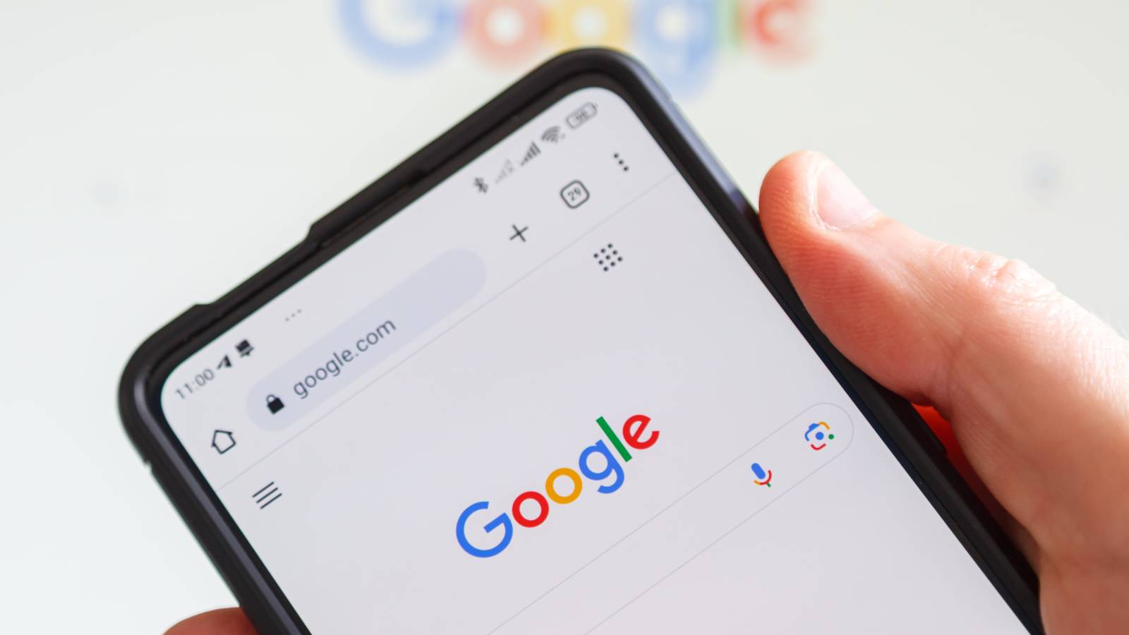 A hand holds a smartphone showing the Google search page, with a blurred Google logo in the background.