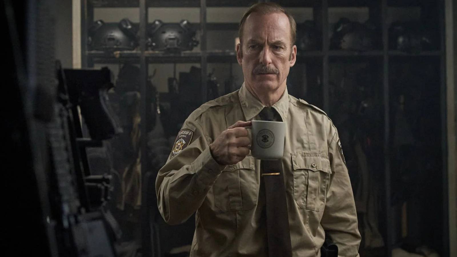 Bob Odenkirk dressed as a sheriff holding a coffee mug against a backdrop of weapons in Normal.