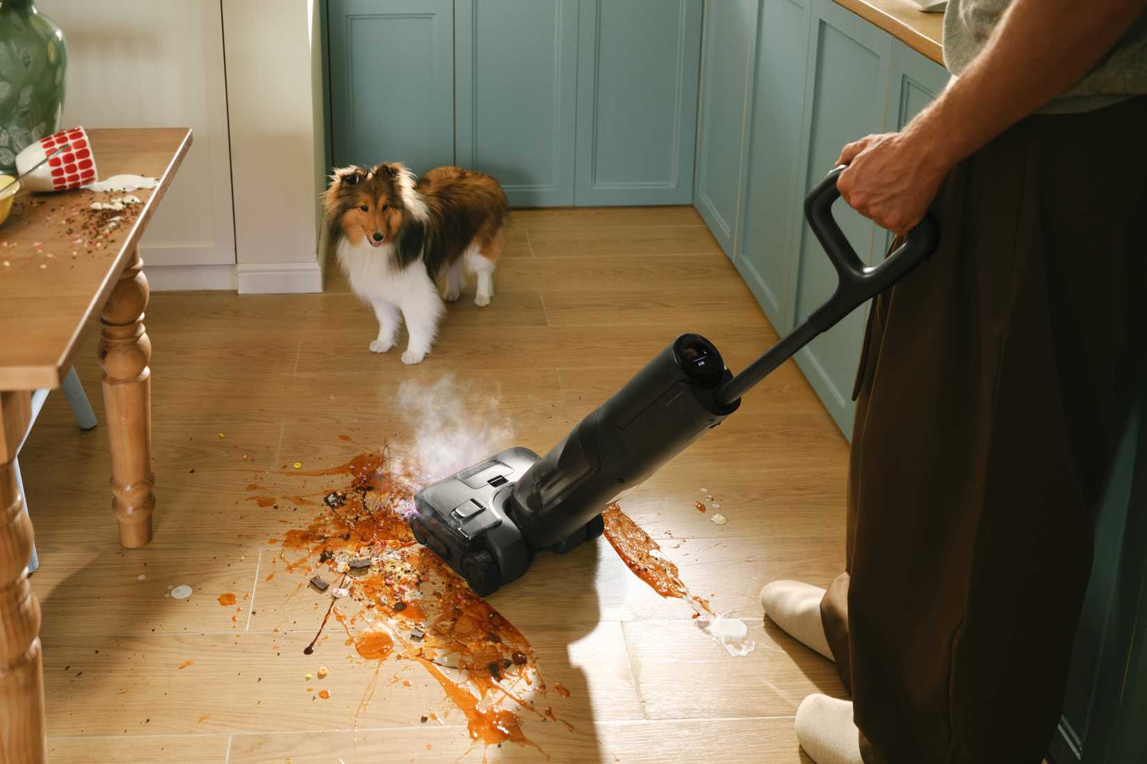 A man using a Roborock F25 Steam to clean up a kitchen mess with a collie in the background