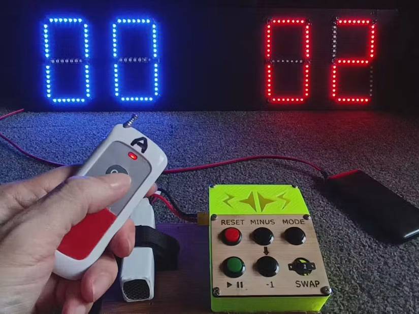 A hand holds a remote beside a button controller and an LED scoreboard showing blue 00 and red 02.