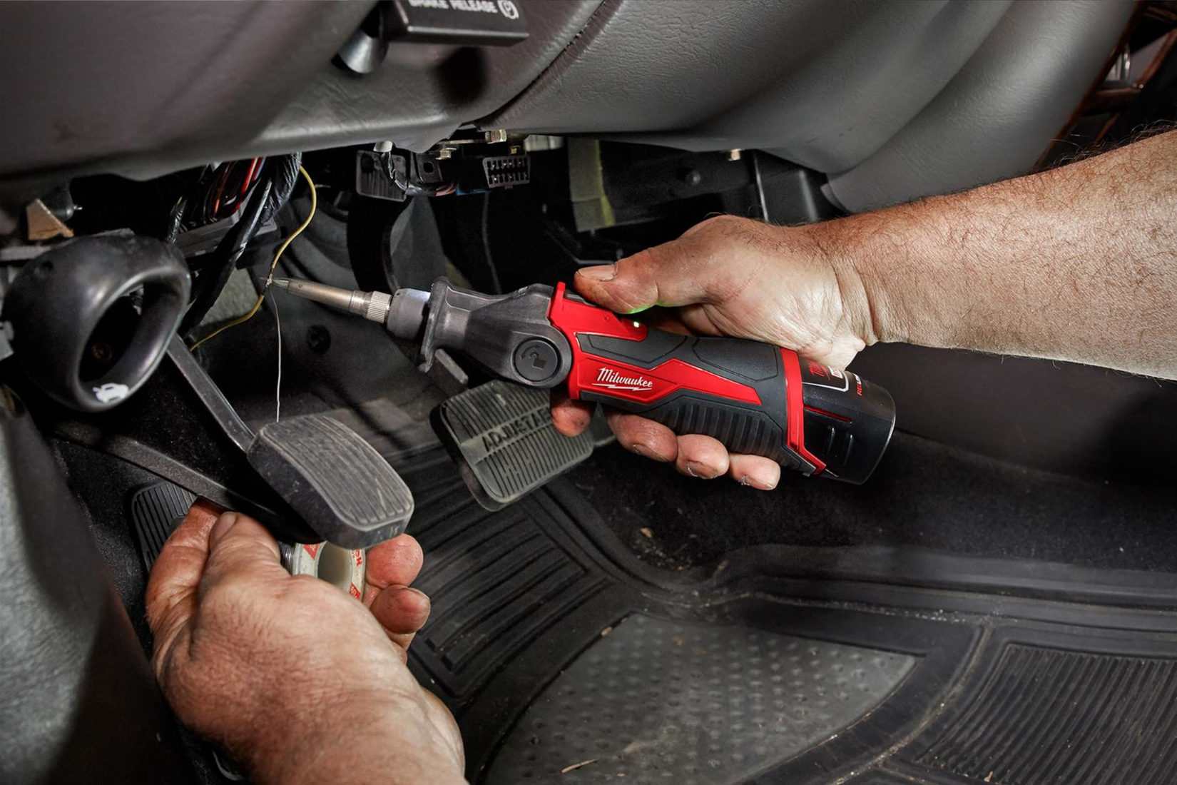 A man using a Milwaukee M12 soldering iron to solder wires under a car's dashboard.