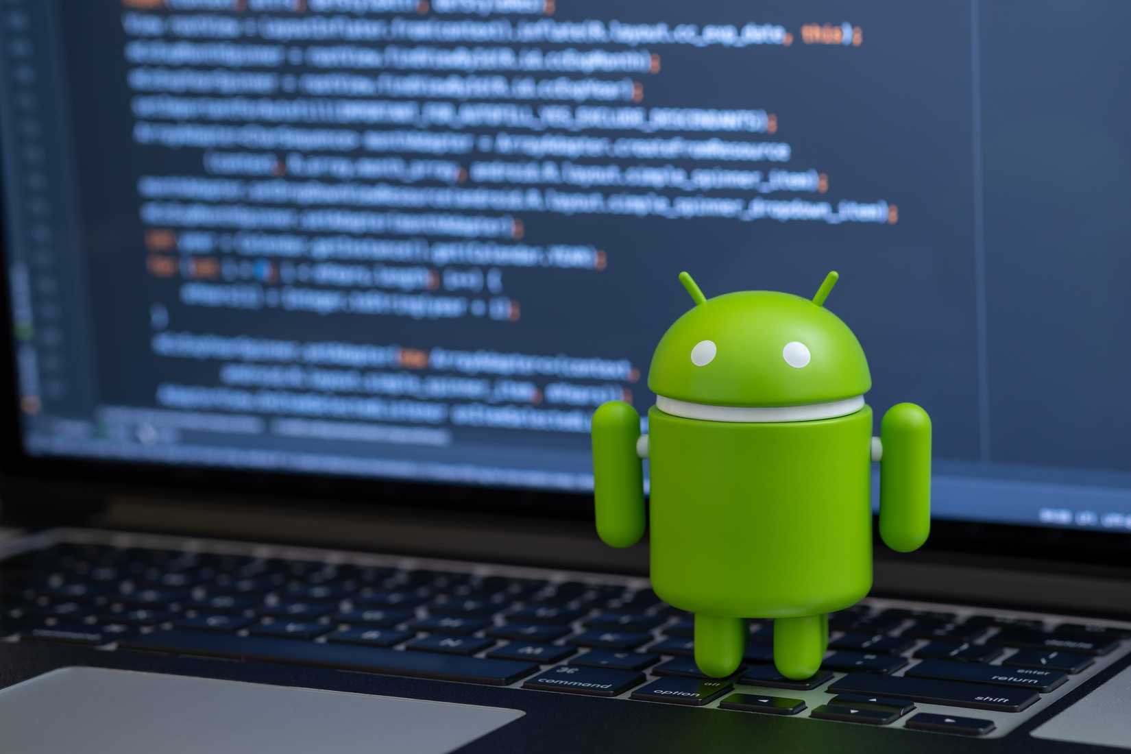 Android robot standing on a laptop keyboard with code running on the laptop screen.