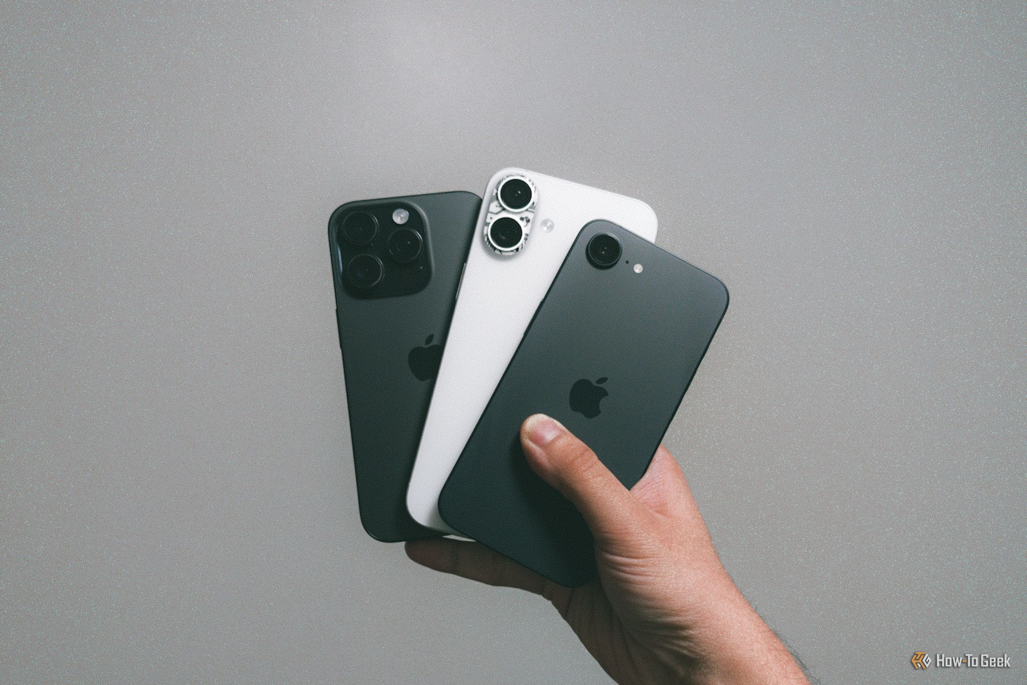 iPhone 16e with iPhone 16 Plus and iPhone 16 Pro in hand against a gray wall.