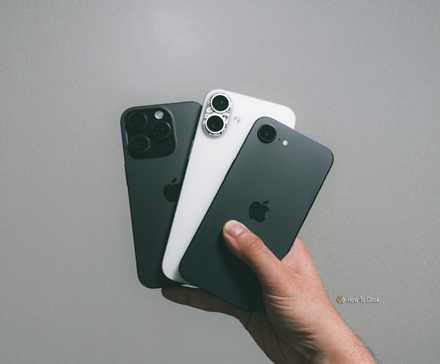 iPhone 16e with iPhone 16 Plus and iPhone 16 Pro in hand against grey wall.