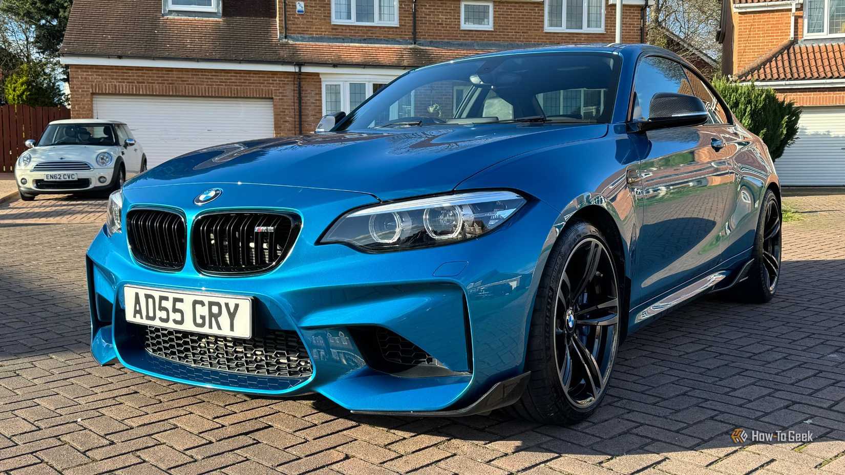 Static front 3/4 shot of a blue 2018 BMW M2 with houses in the background.