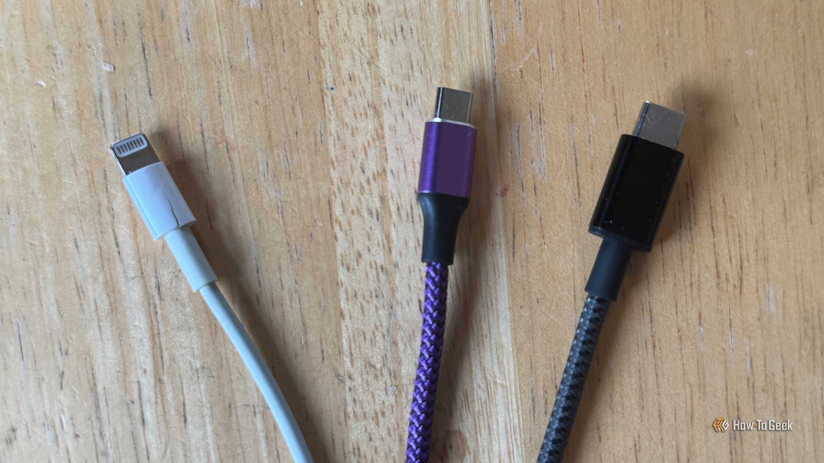 A close up of a white Lightning cable, a purple micro-USB cable and a gray USB-C cable on a table.