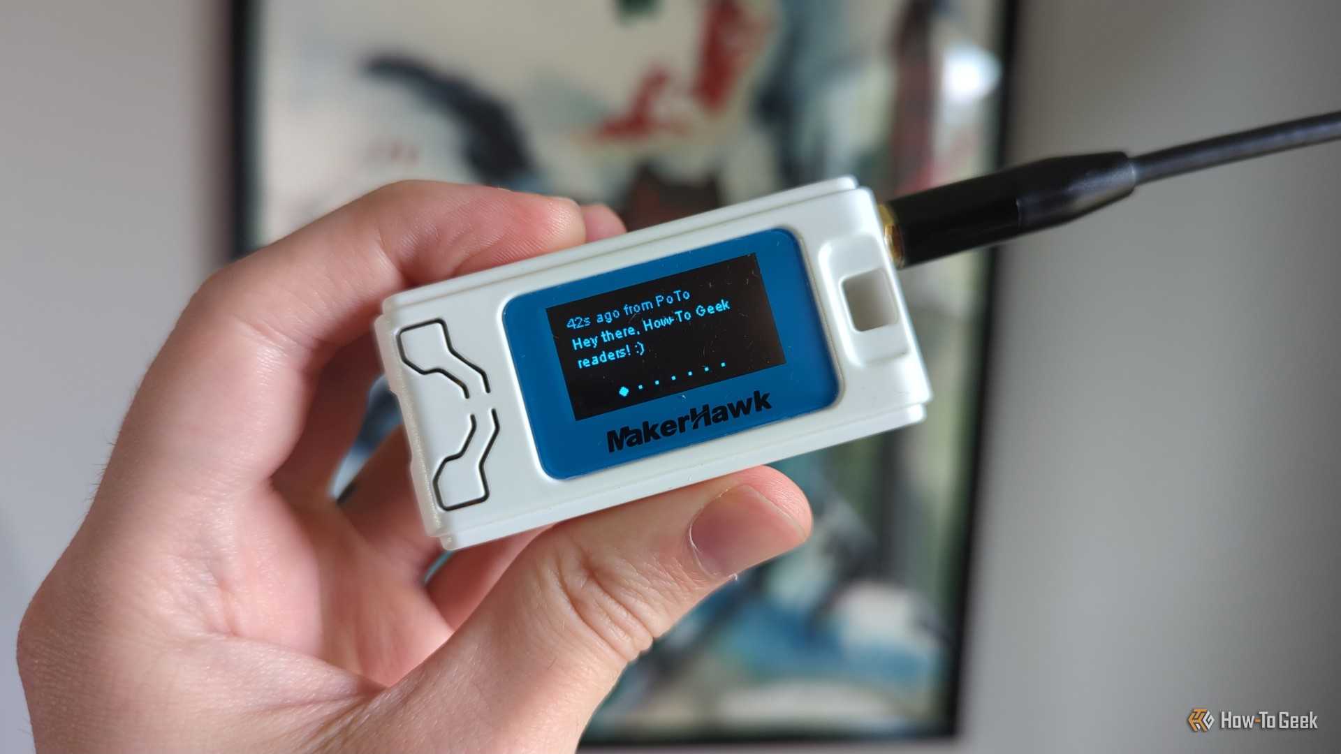 A hand holding a Heltec V3 LoRa radio in a MakerHawk case, with a message on the radio's OLED display reading "Hey there, How-To Geek readers" followed by a smile emote.