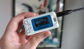 A hand holding a Heltec V3 LoRa radio in a MakerHawk case, with a message on the radio's OLED display reading "Hey there, How-To Geek readers" followed by a smile emote.