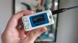 A hand holding a Heltec V3 LoRa radio in a MakerHawk case, with a message on the radio's OLED display reading "Hey there, How-To Geek readers" followed by a smile emote.