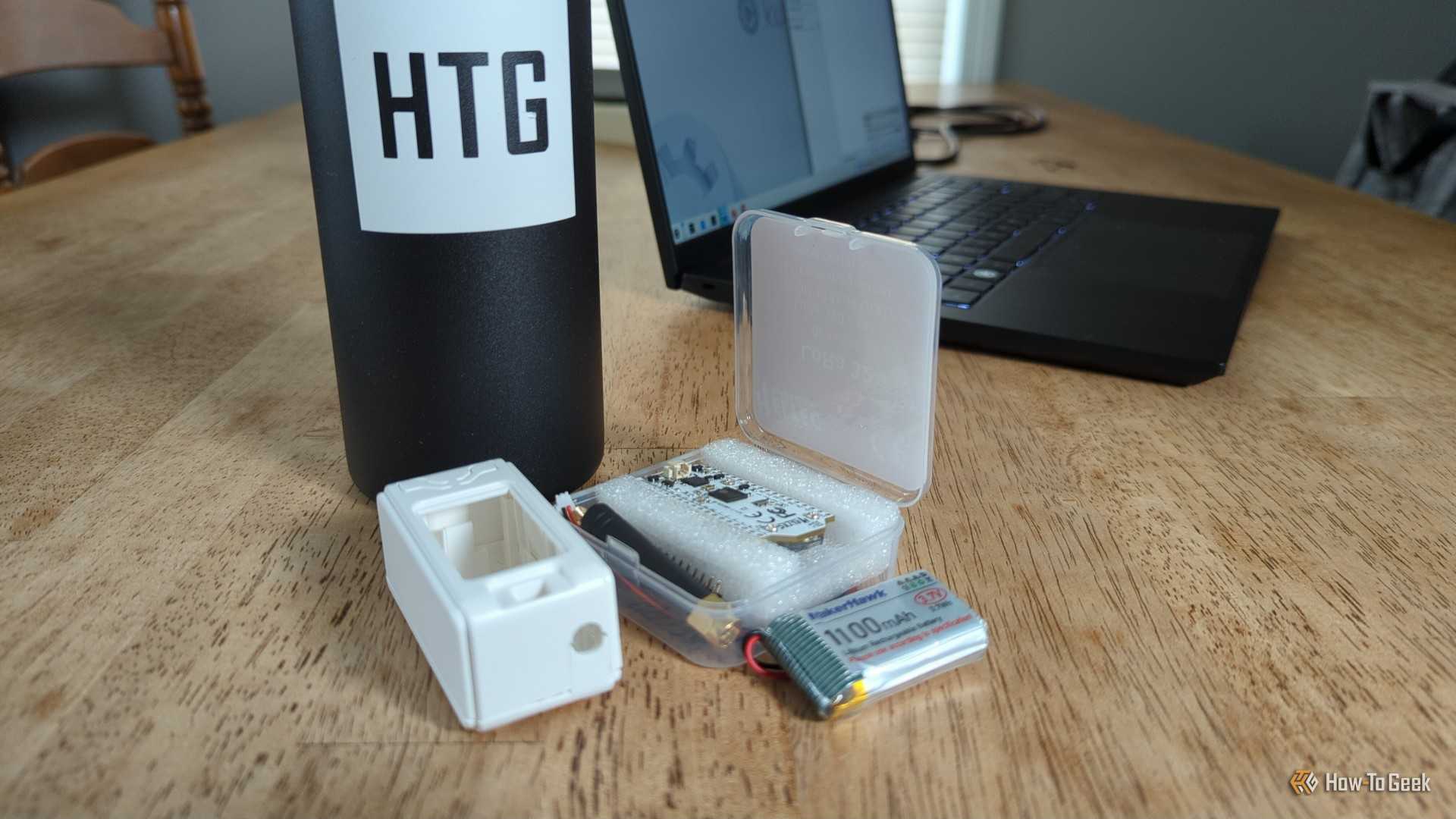 A MakerHawk LoRa radio kit with the board, case, and battery visible in front of a laptop and water bottle with the How-To Geek logo on it.