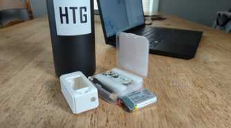 A MakerHawk LoRa radio kit with the board, case, and battery visible in front of a laptop and water bottle with the How-To Geek logo on it.