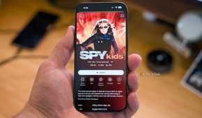 An iPhone 15 Pro held in a hand over a desk with the Spy Kids movie shown on the Plex mobile app.