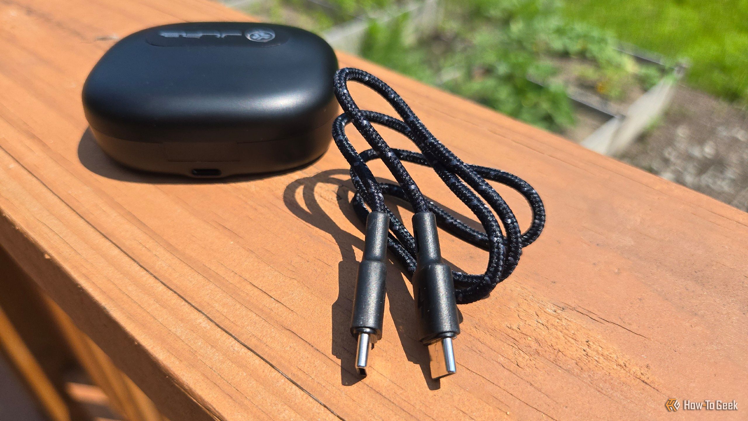 Included USB-C cable next to the case for JLab Epic Open Sport on a deck railing.
