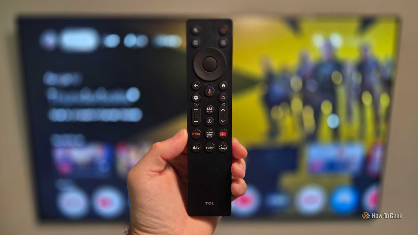 A hand holds the remote control for a TCL QM6K TV. 