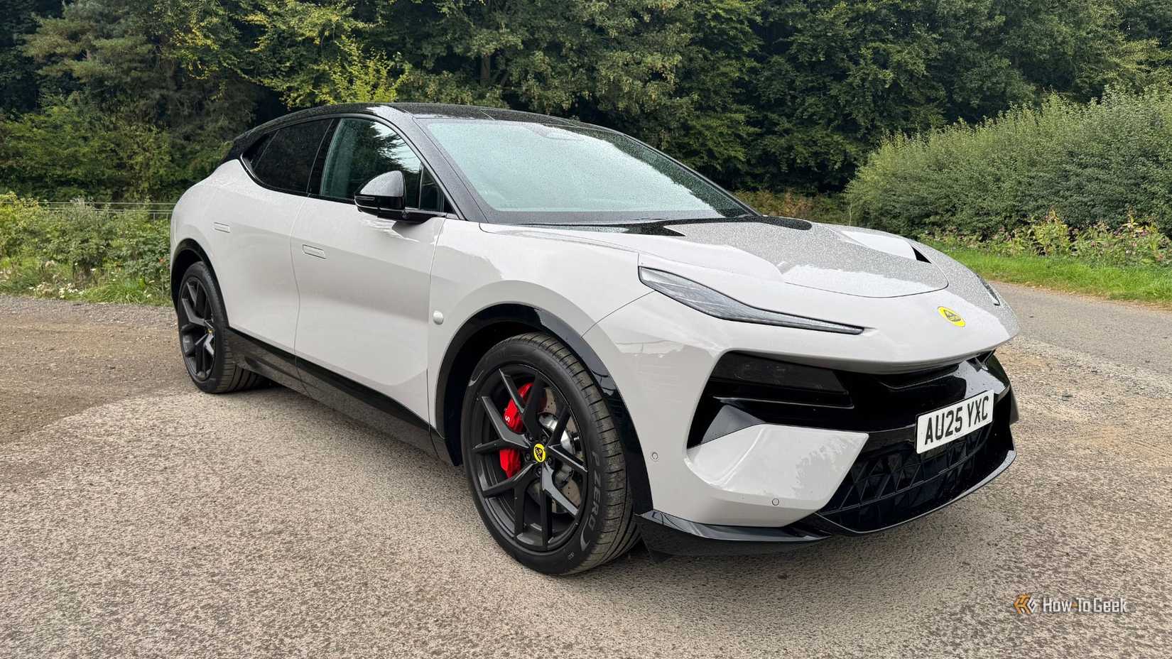 Frente estática 3/4 de um tiro de um cinza 2025 Lotus Eletre 600 Sport SE estacionado em cascalho com árvores ao fundo.