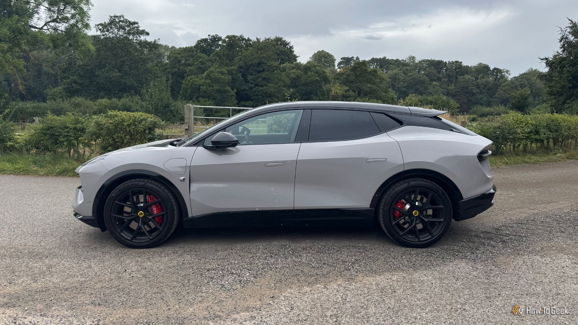 Toma estática de perfil lateral de un Lotus Eletre 600 Sport SE gris 2025 estacionado sobre grava con árboles al fondo.
