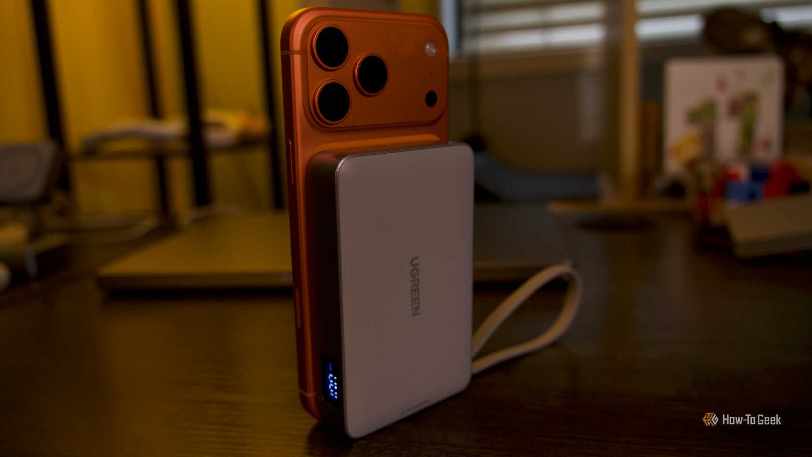 O Power Bank Ugreen MagFlow 10000mAh 25W conectado a um iPhone 17 Pro Max laranja em pé sobre uma mesa.
