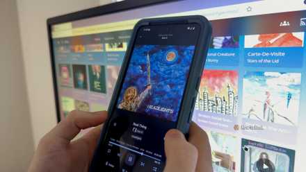 A person's hands holding an Anrdoid phone with the Finamp app in front of a computer screen.