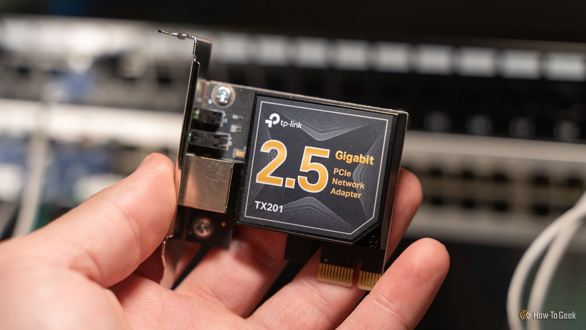 A TP-Link 2.5 gigabit PCIe networking adapter held by a person with network switches in the background.