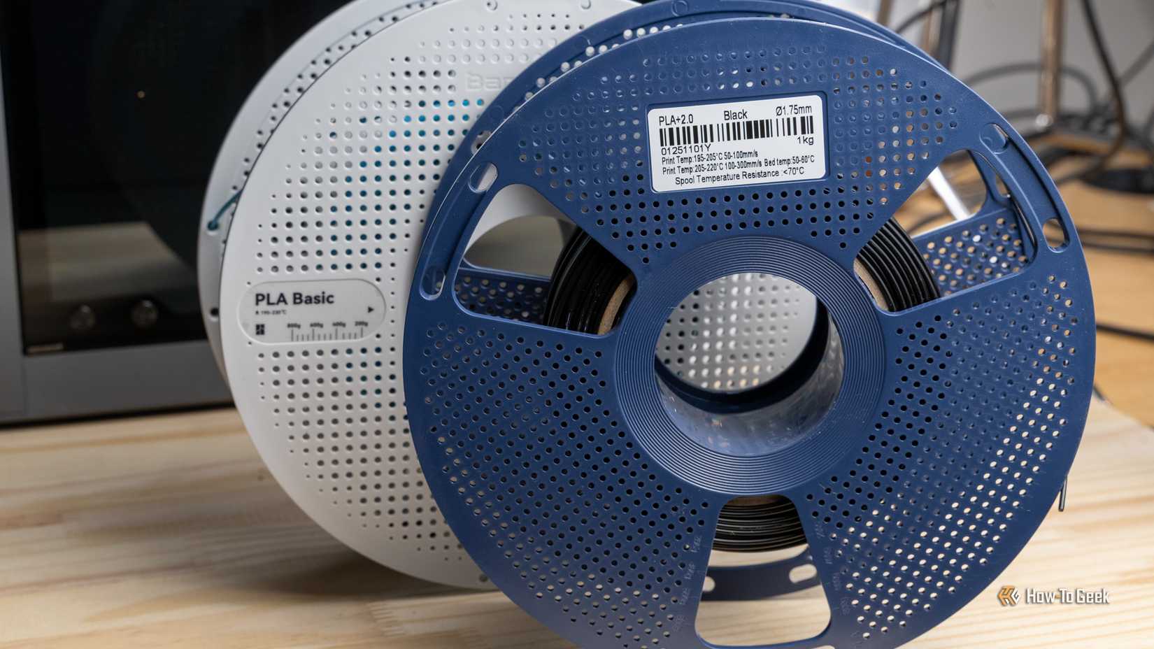 A blue spool of black PLA+ 2.0 filament in front of a white Bambu Lab PLA Basic spool on a wooden desk.