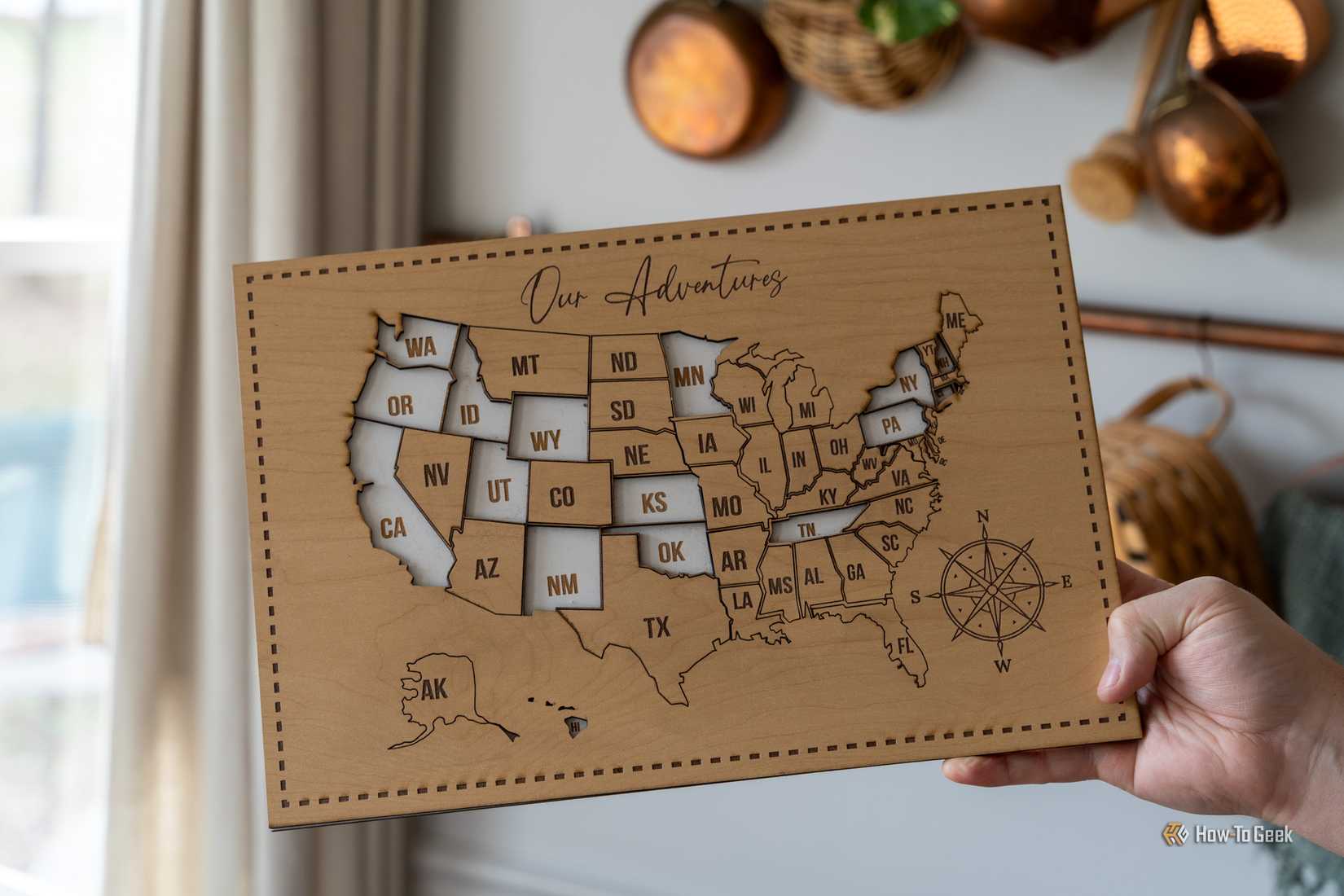 A hand holding a completed laser-cut wooden US map titled Our Adventures with a compass rose.