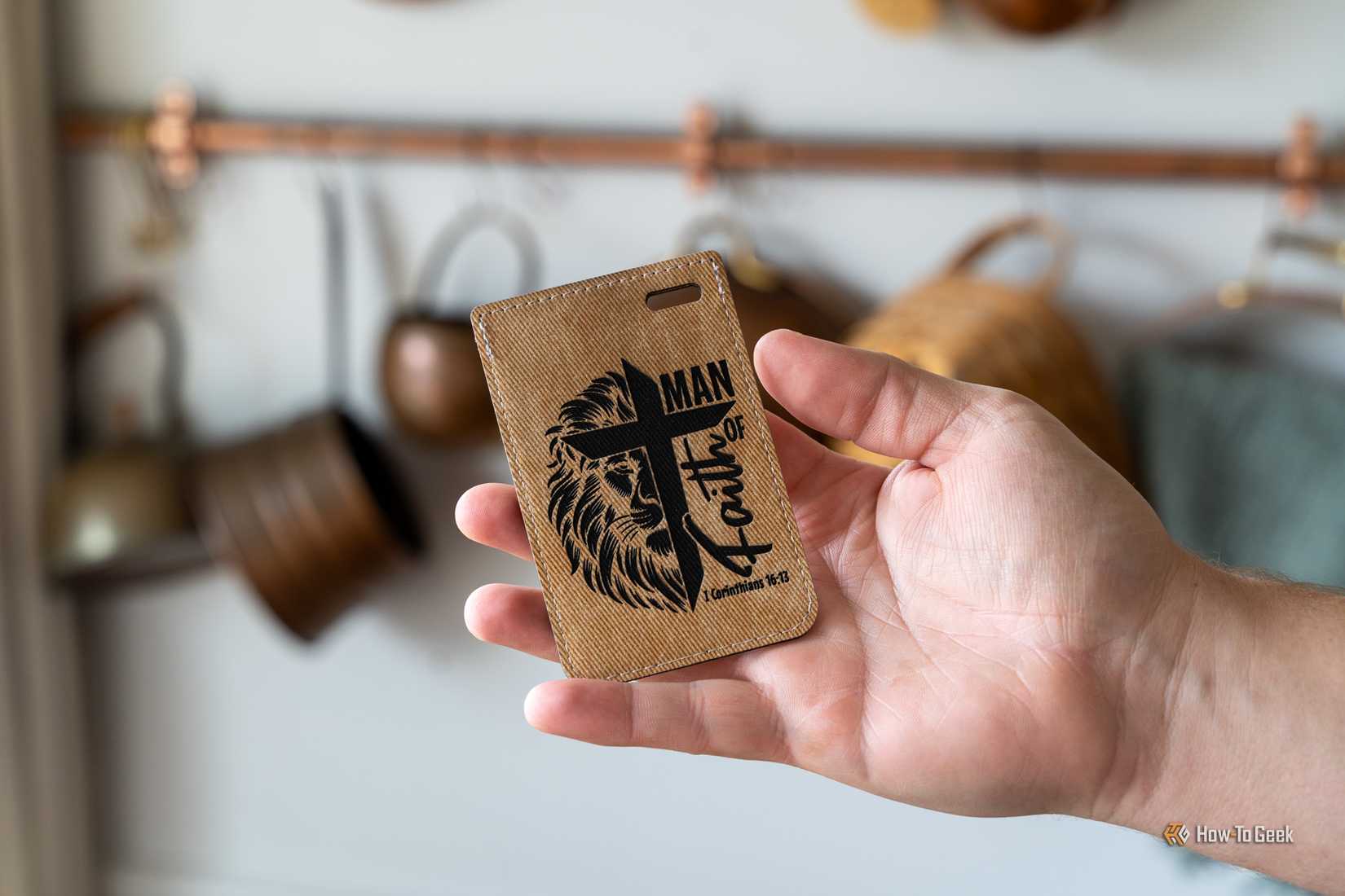 A hand holding a laser-engraved leather luggage tag with a Man of Faith lion and cross design.