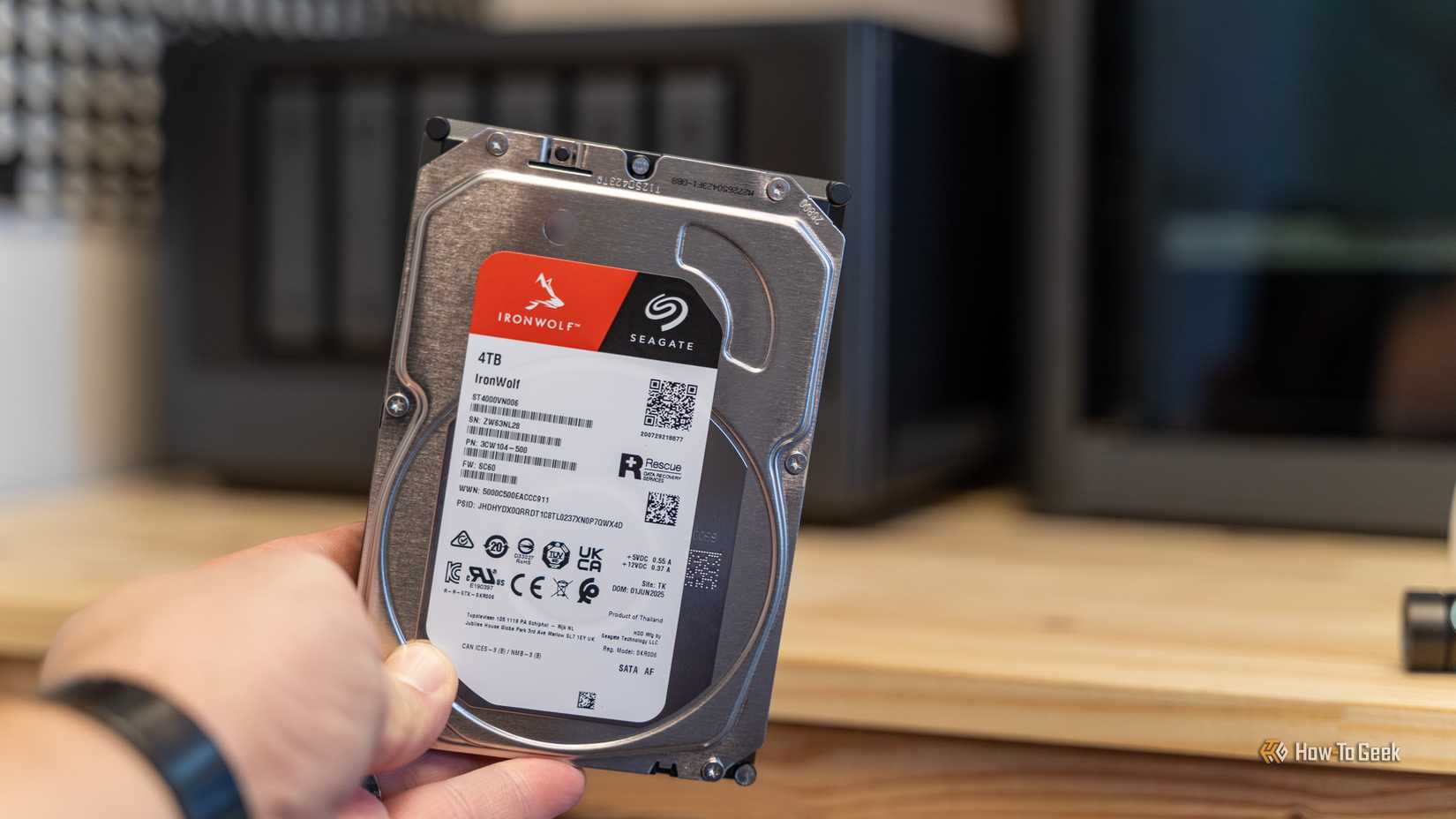 A hand holding a Seagate IronWolf 4TB hard drive with the Ugreen iDX6011 Pro NAS in the background.
