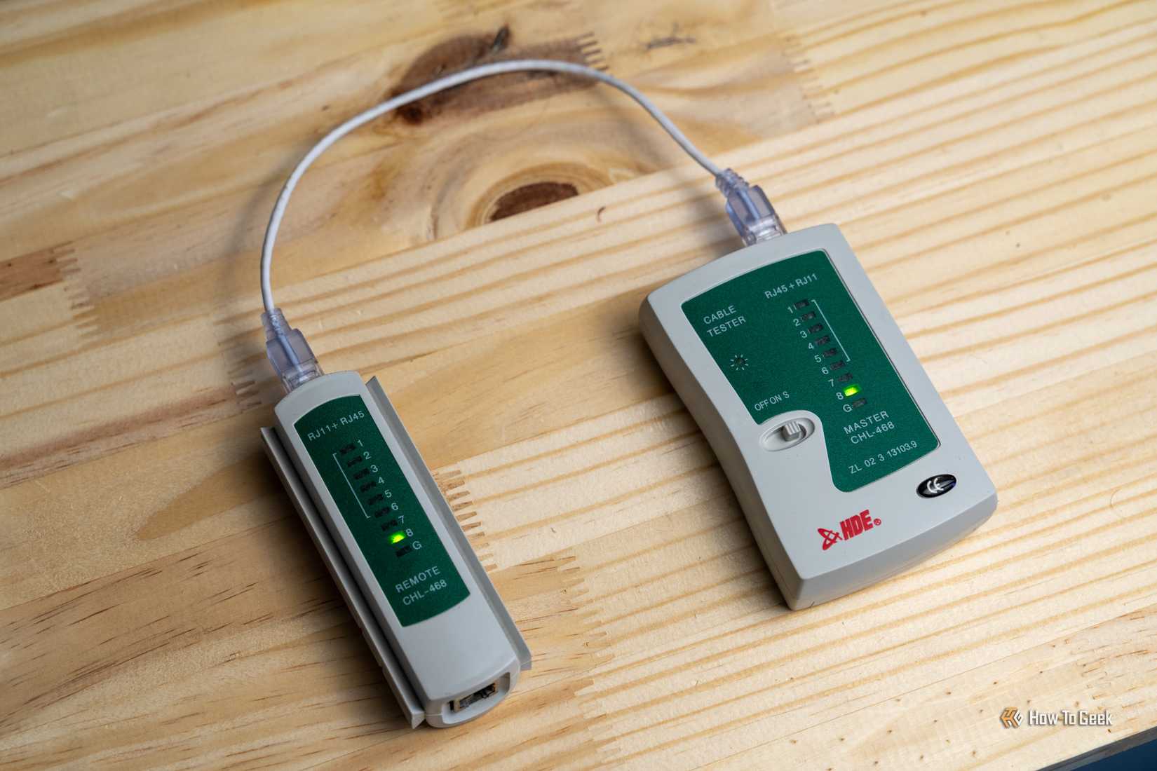 A gray network cable tester connected with a short Ethernet cable on a wooden workbench.
