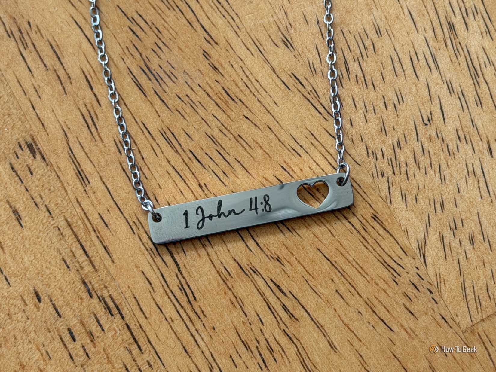 A silver bar necklace engraved with 1 John 4:8 and a heart-shaped cutout lies on a wooden table.
