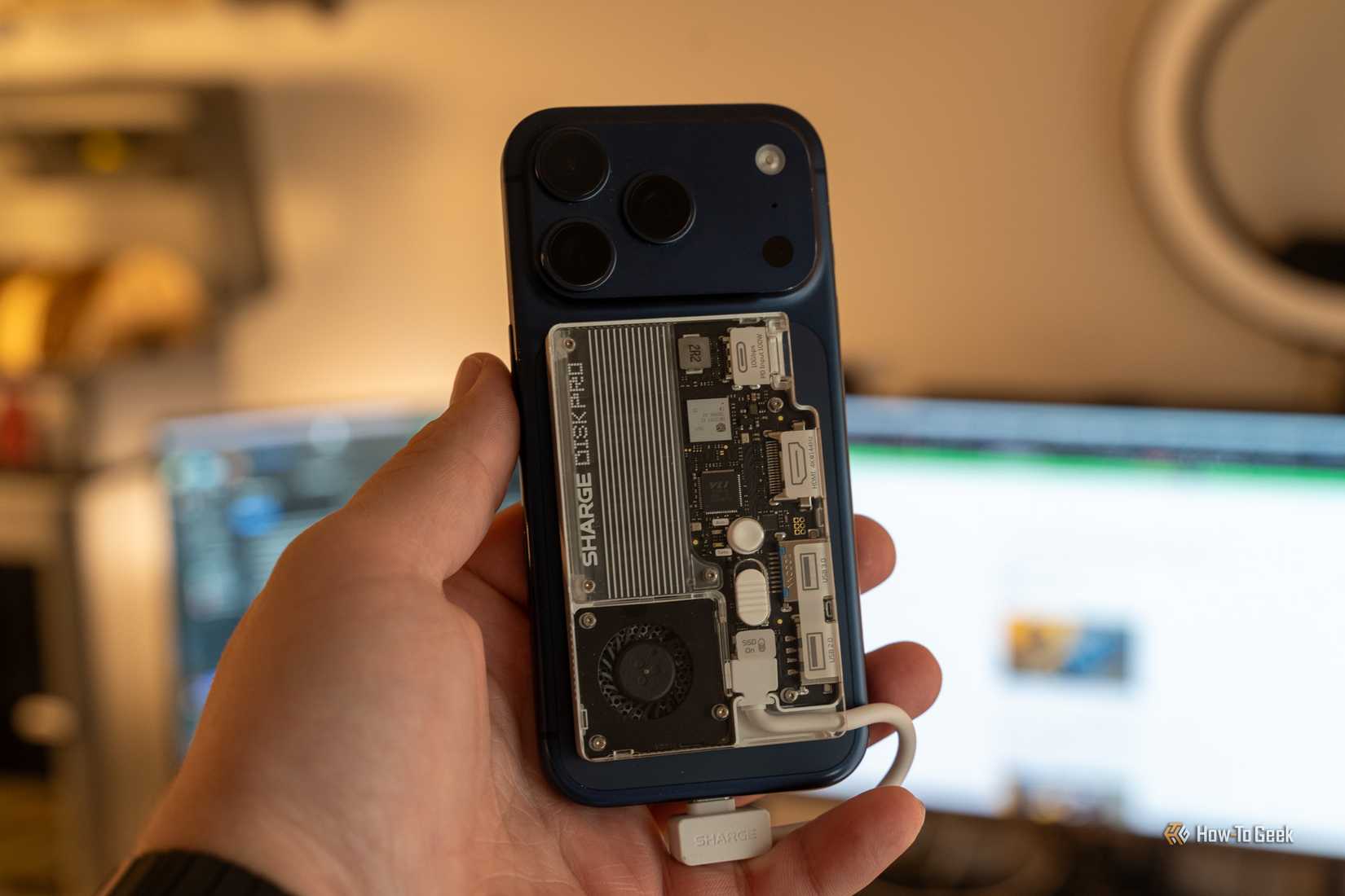A hand holding the Sharge Disk Pro attached to the back of a blue iPhone 17 Pro in front of a blurred monitor.