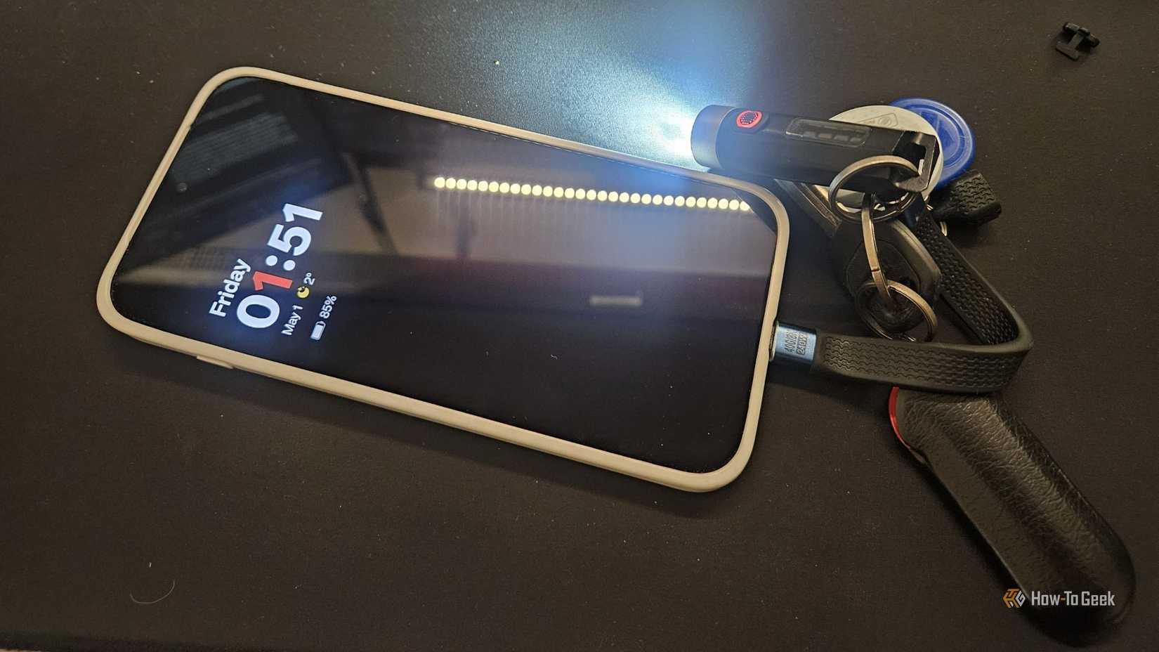 A phone with a USB-C cable plugged into it and a keychain flashlight running and a small cat hair next to the phone.