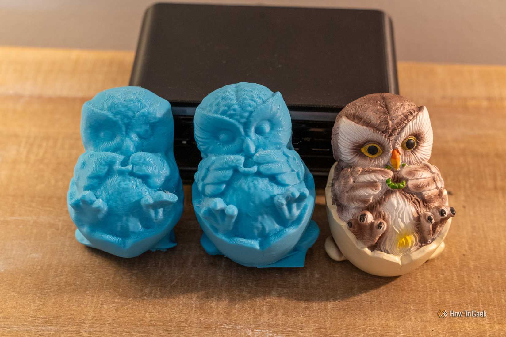 Two blue 3D-printed owl scans beside the original painted owl figurine with an iPhone behind them.