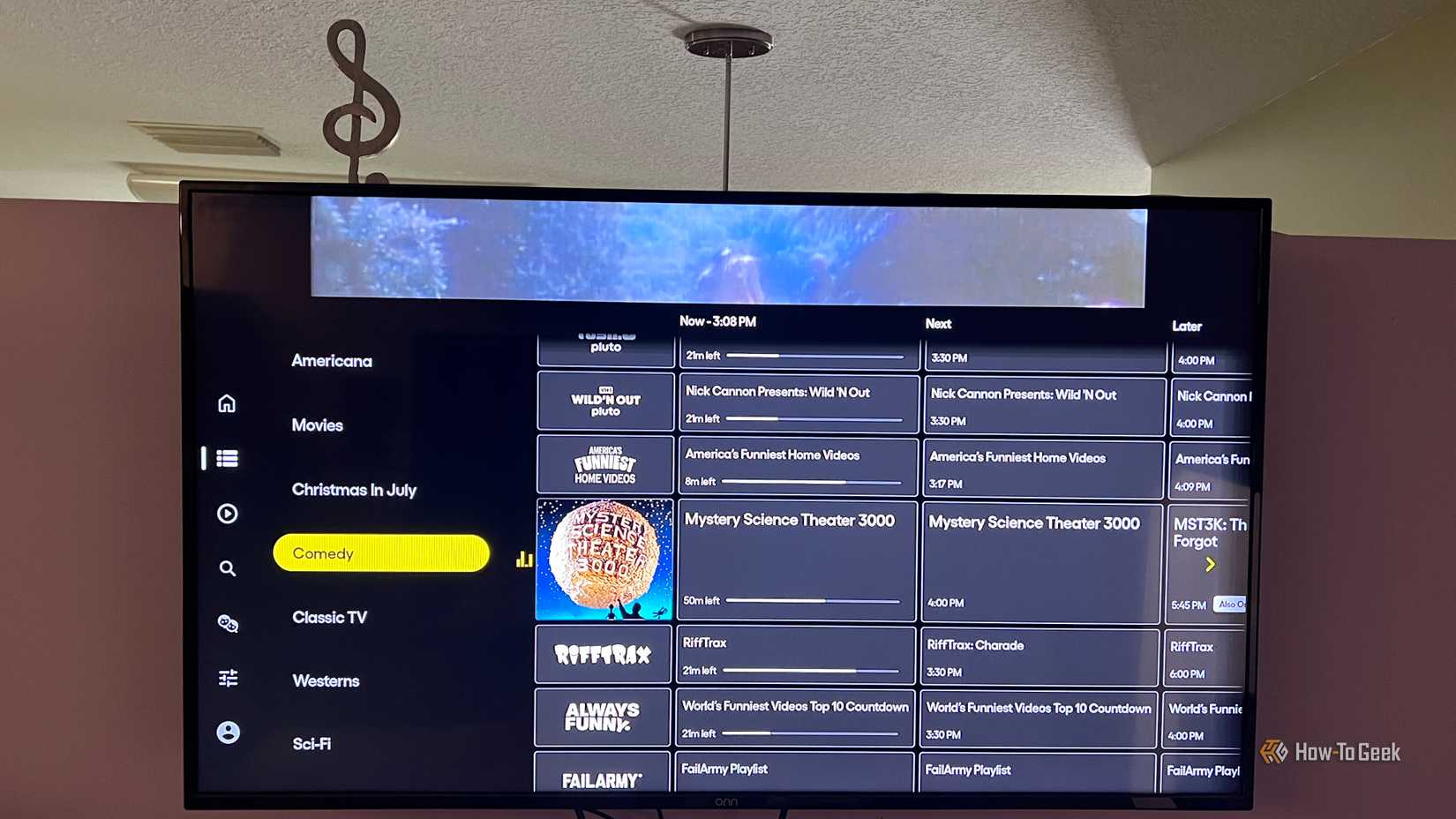 An image of a TV hanging on a wall with the streaming app Pluto TV running. The Channel guide is open and the Comedy category is displaying shows in that category.