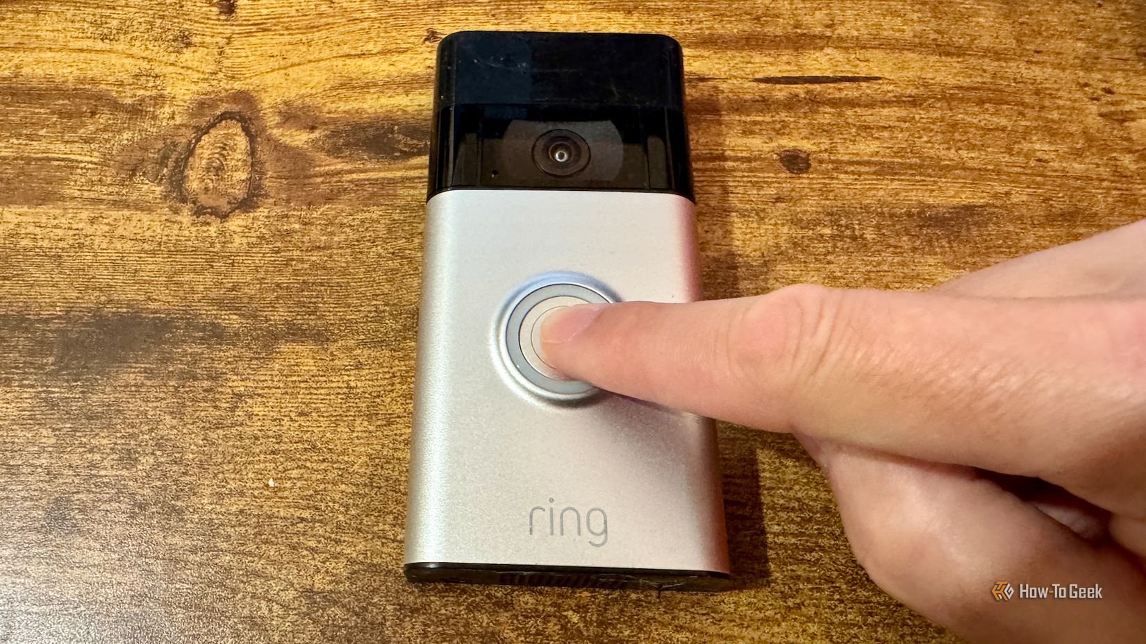 A finger pressed the ring button of a Ring video doorbell on a desk.
