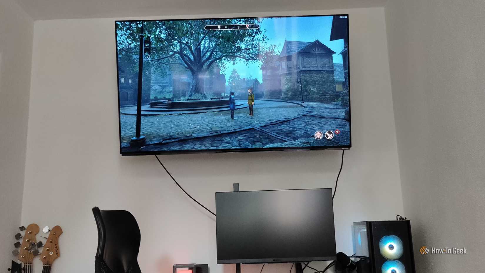 A TV mounted to the wall showing a video game above a desktop PC.