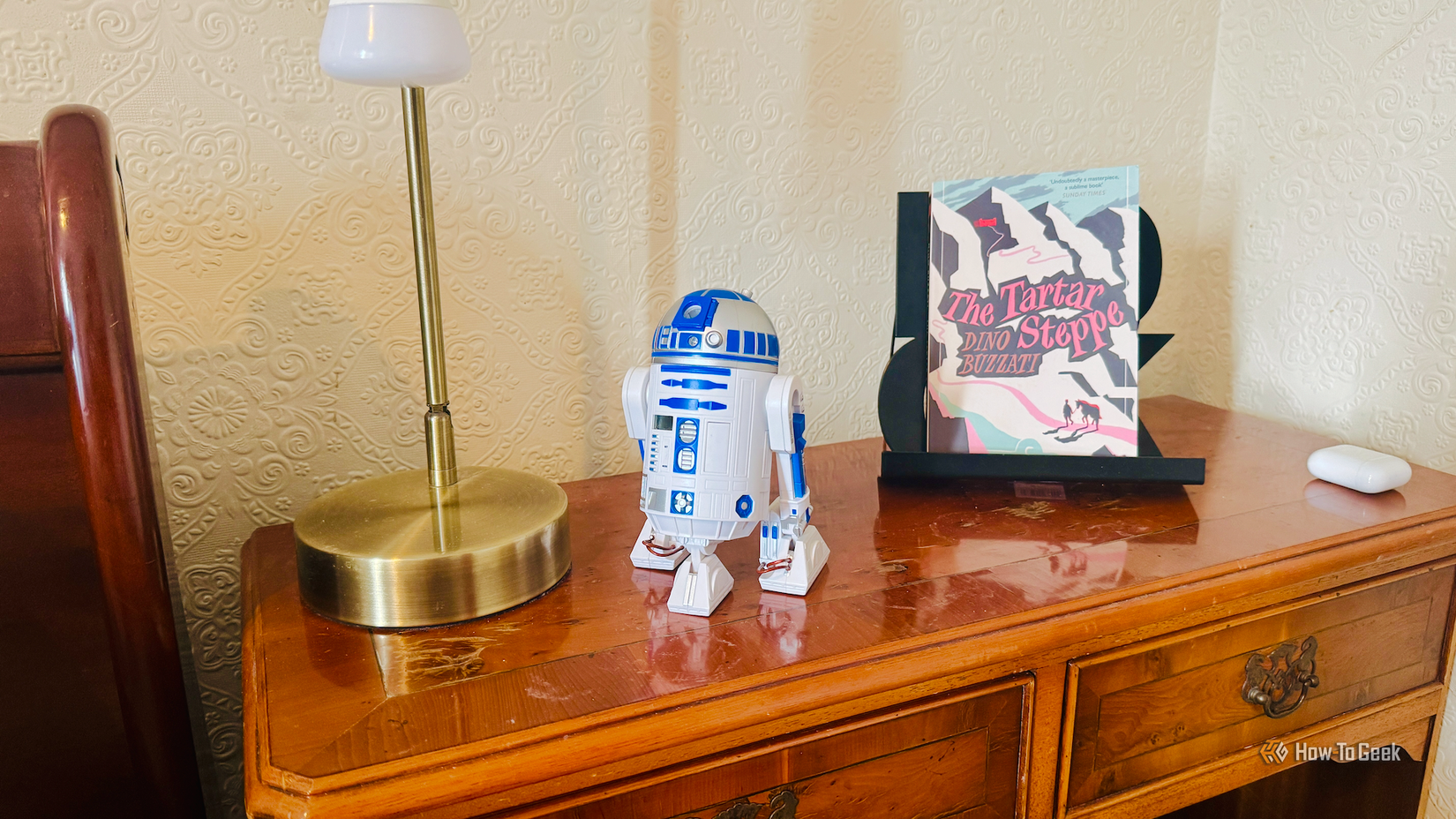 An R2D2 alarm clock on a bedside table next to a lamp, an awesome book, and some AirPods.