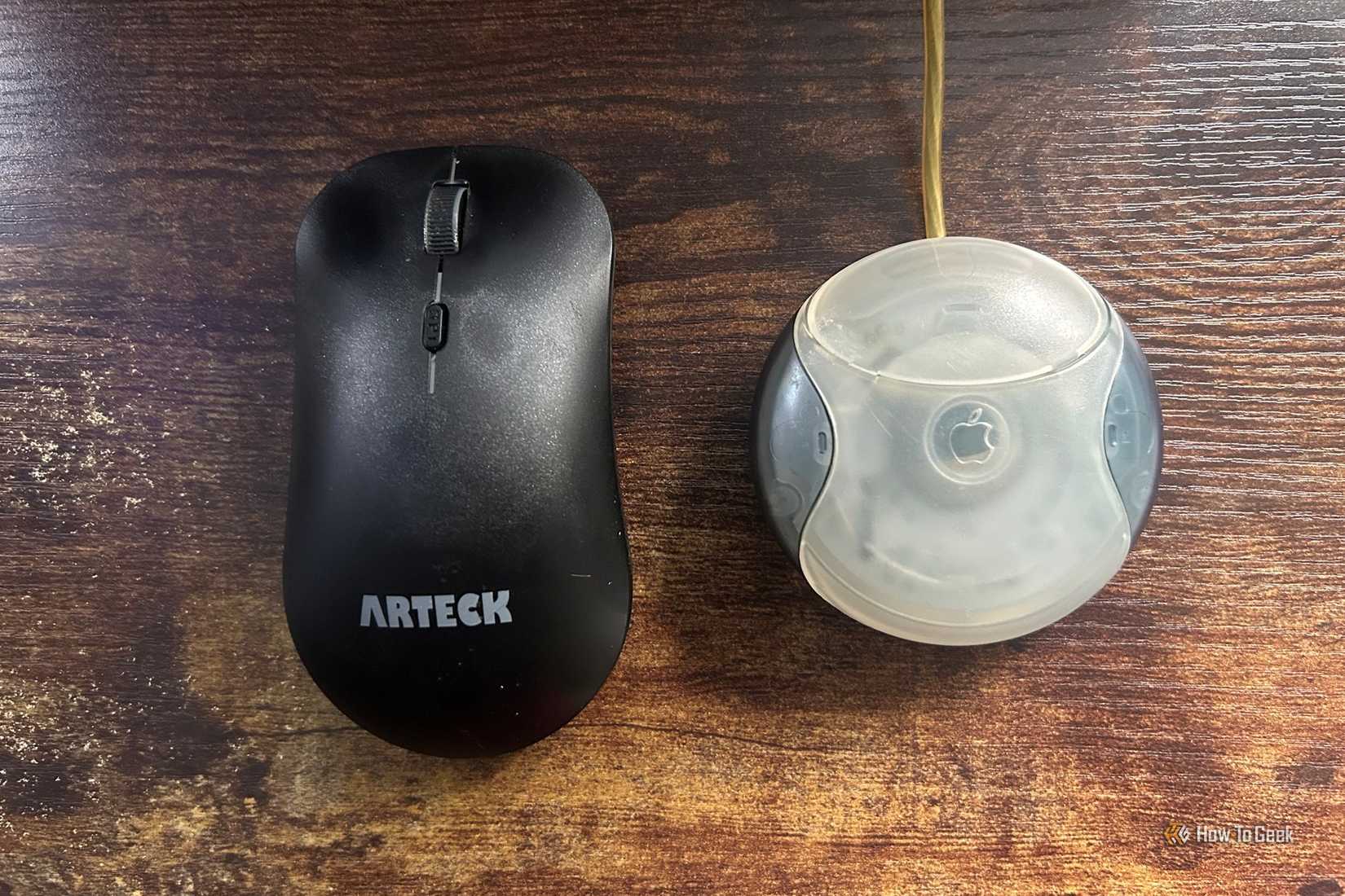 A black Arteck mouse placed next to a vintage Apple USB hockey puck mouse on a wooden surface.