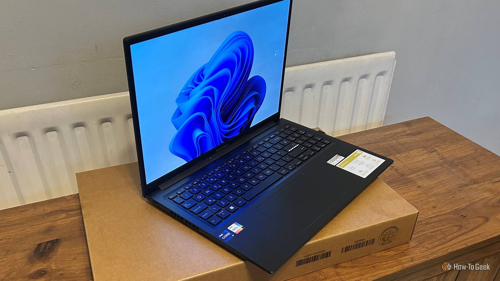 Windows 11 desktop wallpaper displays on an open ASUS Vivobook laptop resting on its cardboard box.