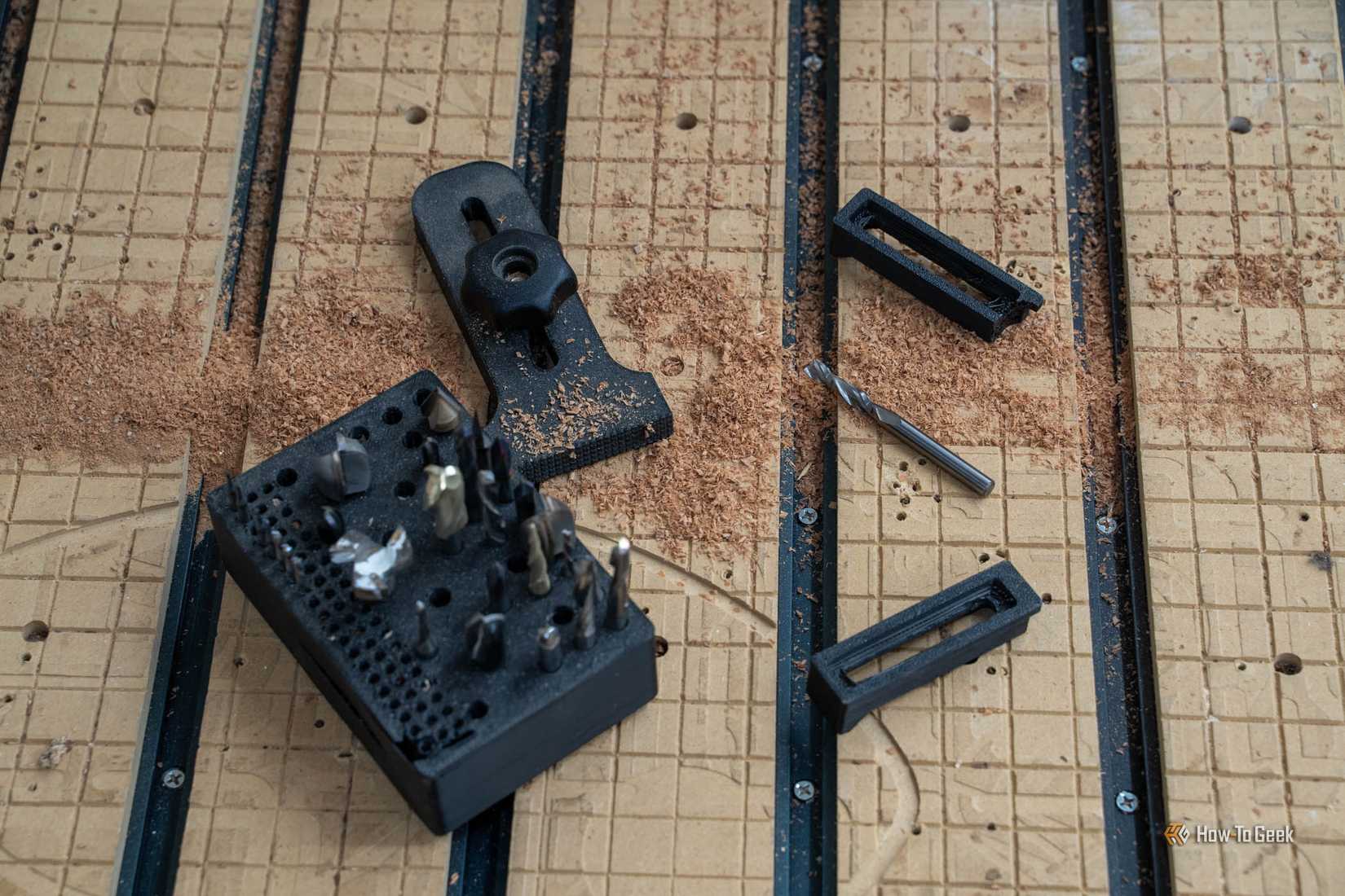 A black bit holder full of router bits beside a knob hold-down clamp and two printed CNC clamps.