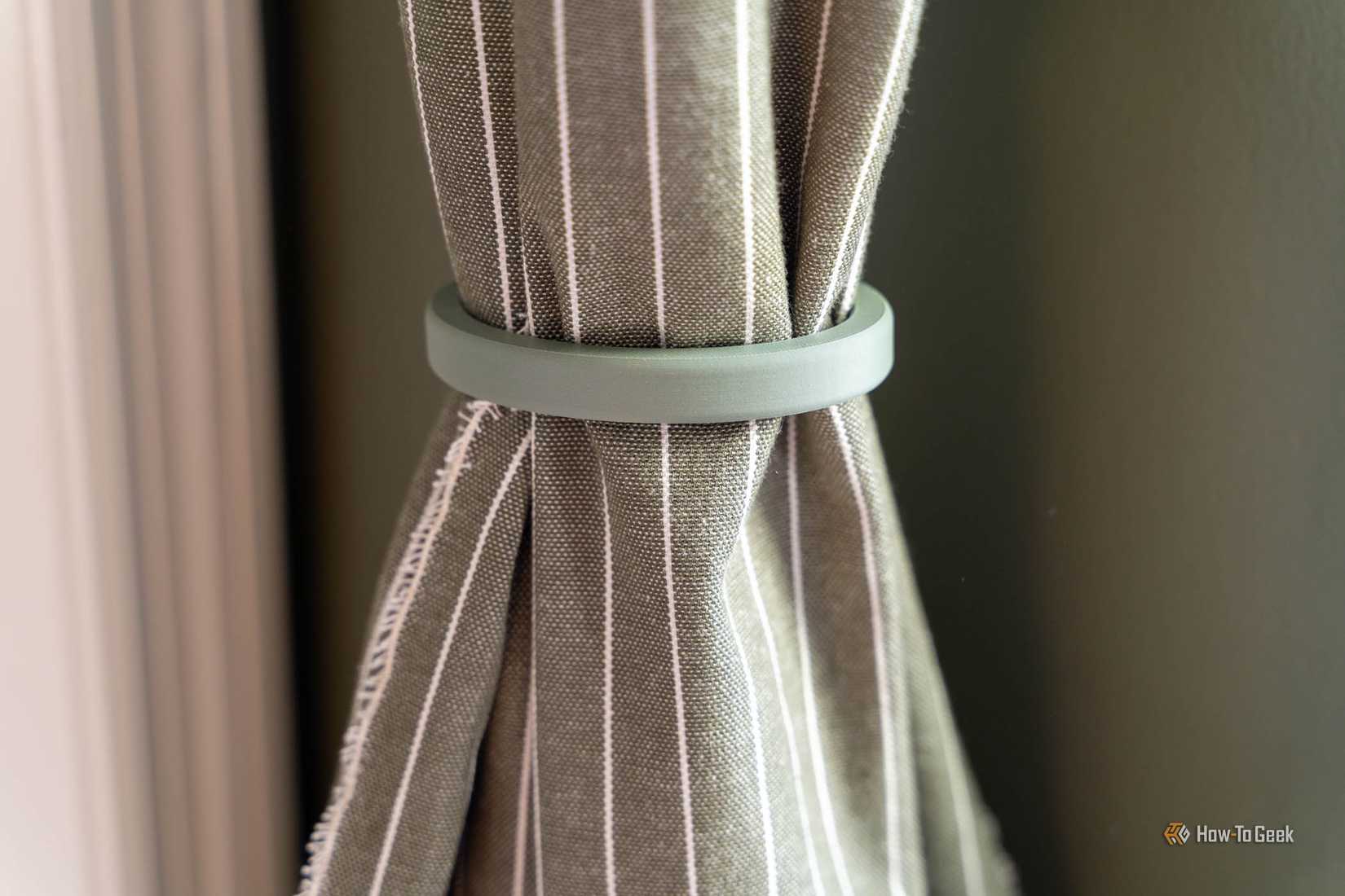 Green 3D-printed curtain tie ring gathering a striped olive curtain without any wall hardware.
