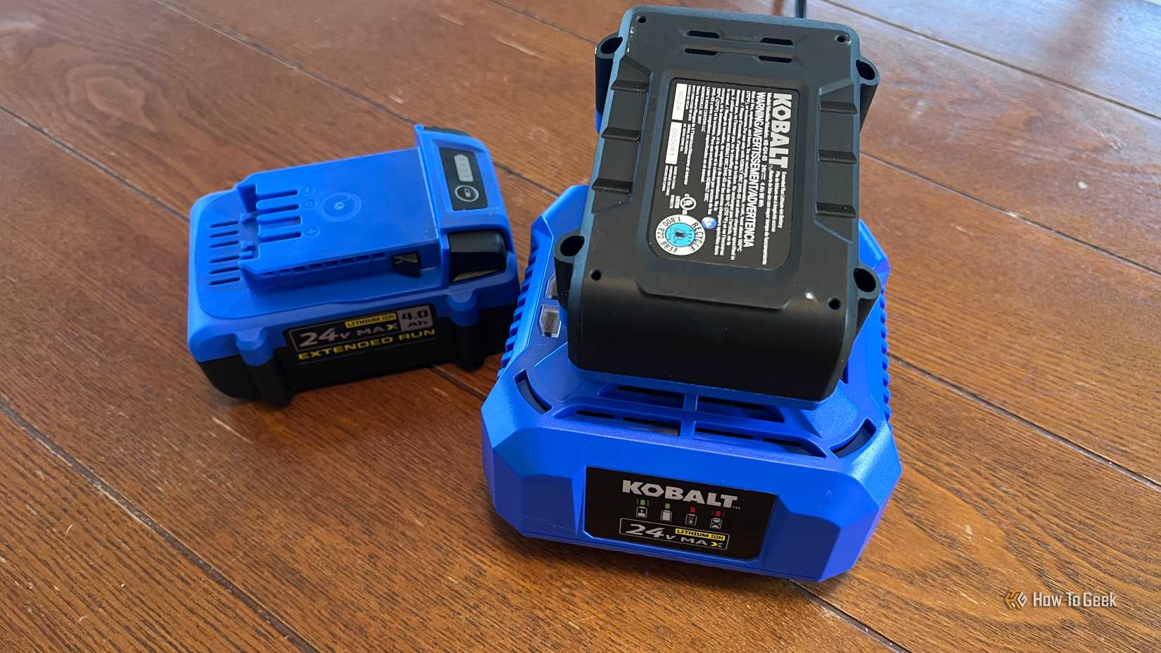 Two Kobalt 24V lithium-ion batteries are displayed on a wooden floor, with one battery connected to its matching charger.