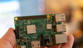Close-up of a Raspberry Pi 3 Model B held in a hand, showing the V1.2 board label, GPIO header, and heatsinks.