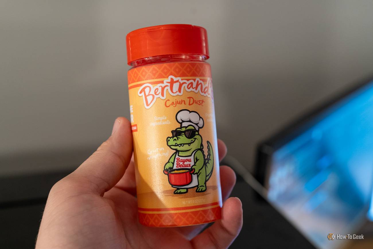 A hand holds a seasoning jar with a red lid and a colorful UV-printed Cajun Dust label featuring an alligator.