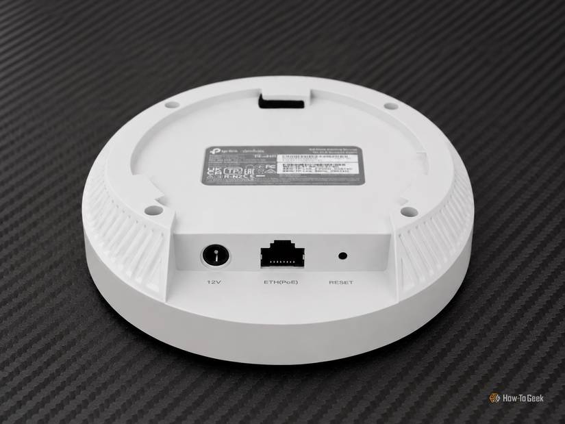 White TP-Link Omada access point shown from the underside on a carbon-fiber mat, with 12V, Ethernet PoE, and reset ports visible.